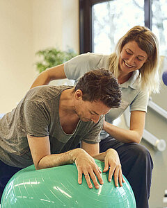 Ein Medical Park Patient stützt sich auf einem Gymnastikball ab während die Therapeutin ihm dabei unterstützt.