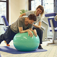 Ein Medical Park Patient stützt sich auf einem Gymnastikball ab während die Therapeutin ihm dabei unterstützt.