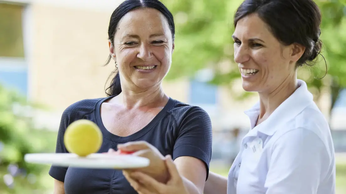 Eine Therapeutin unterstützt eine Medical Park Patientin einen Tennisball mit einer Tenniskelle zu jonglieren. 